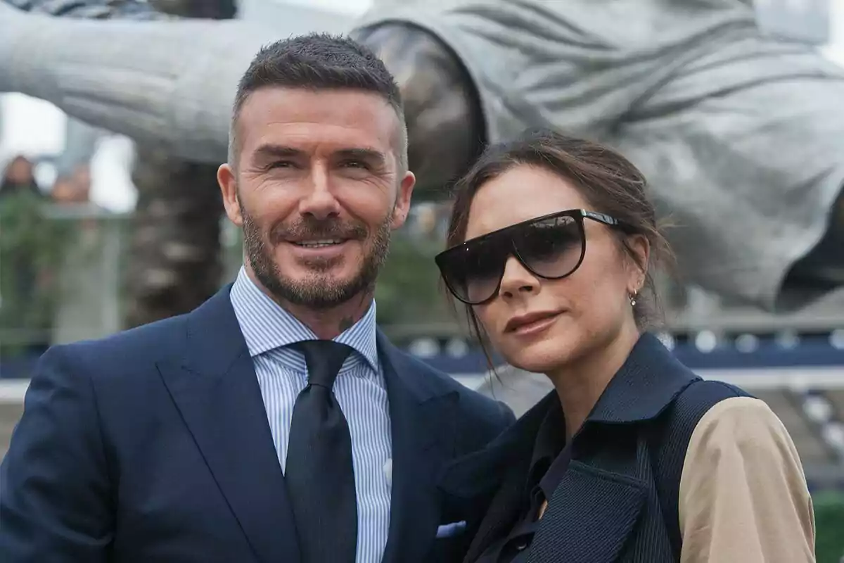 David Beckham and Victoria Beckham posing together, he is wearing a suit and tie, she is wearing large sunglasses.