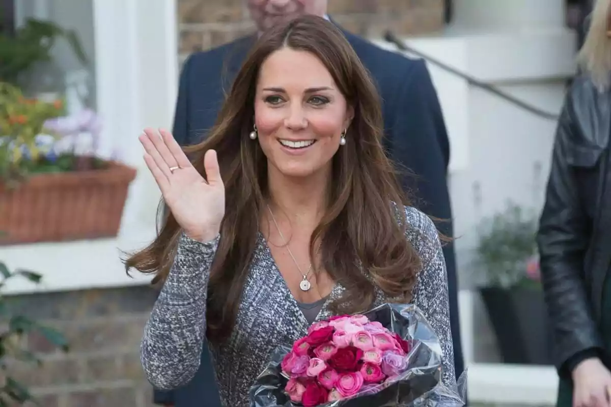 A smiling woman with long brown hair waves while holding a bouquet of pink flowers.
