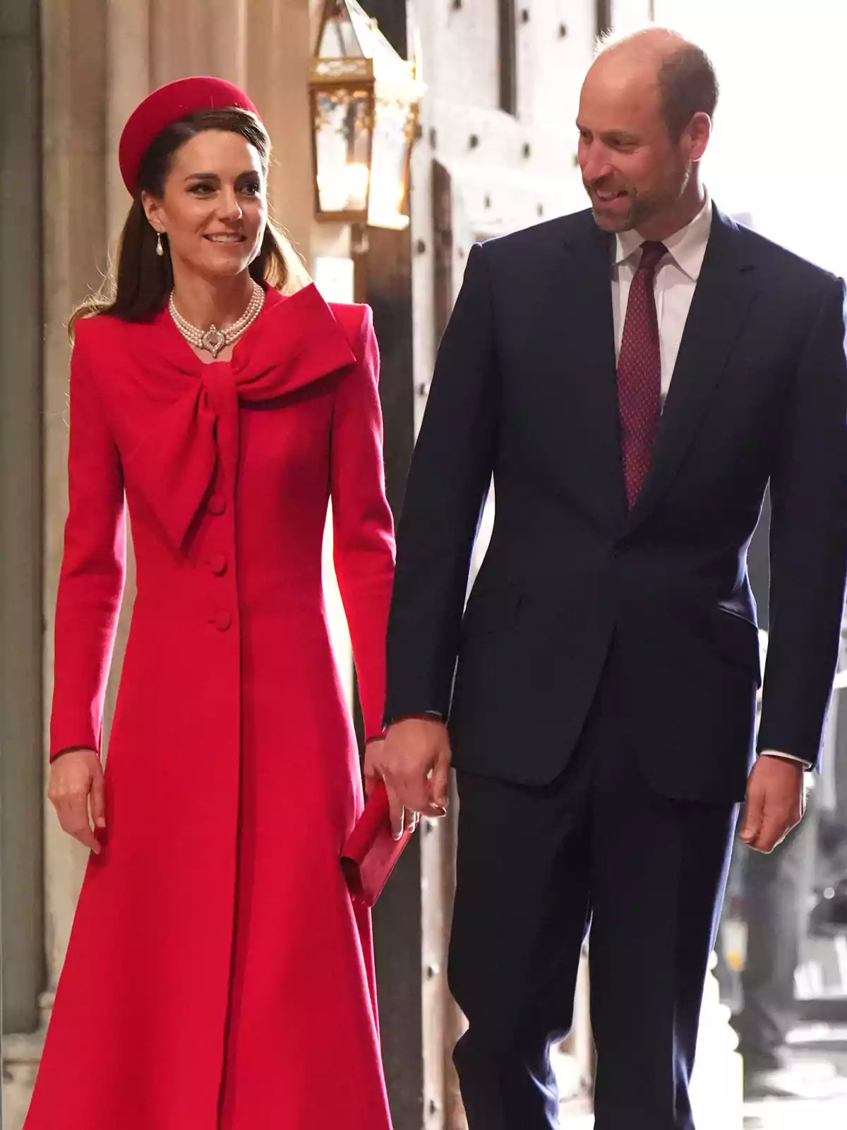 Kate Middleton and Prince William elegantly dressed walk hand in hand at a formal event, she wears a red dress with a matching hat and he wears a dark suit with a tie.