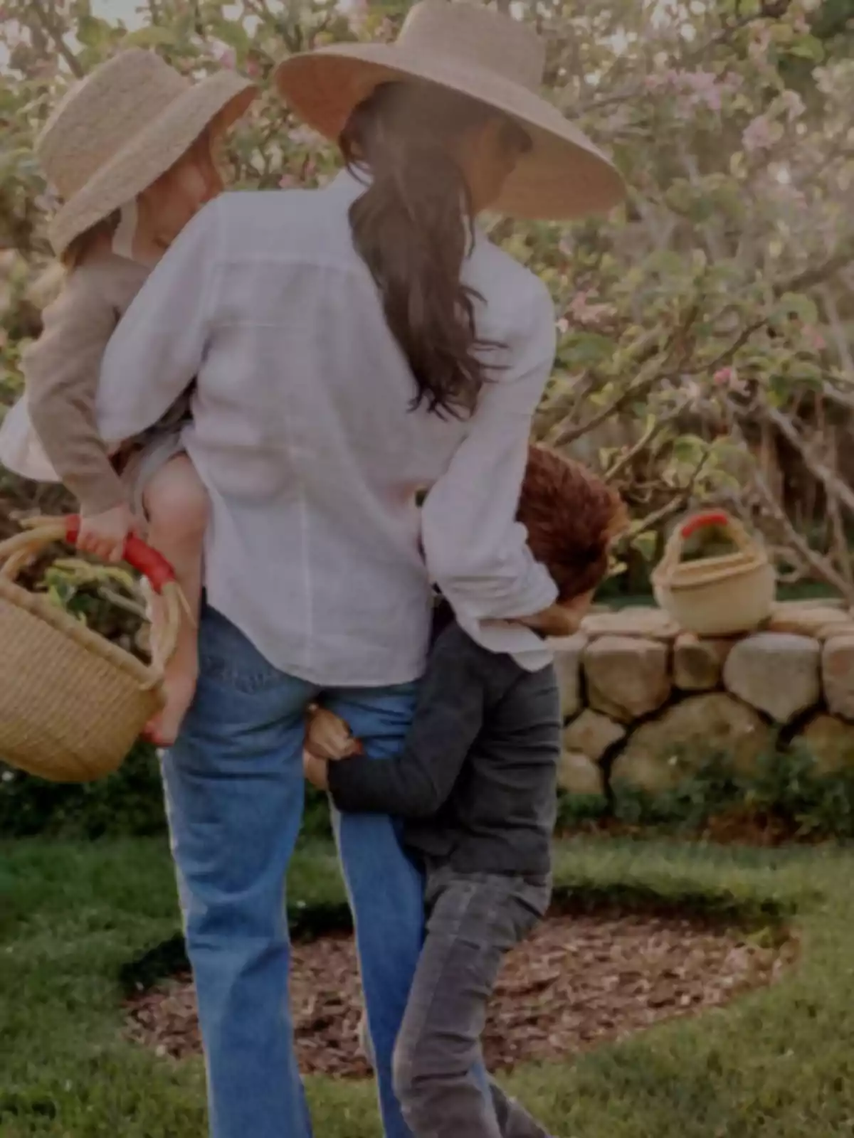 A person with a straw hat carries a child in their arms while another child hugs their leg in a garden.