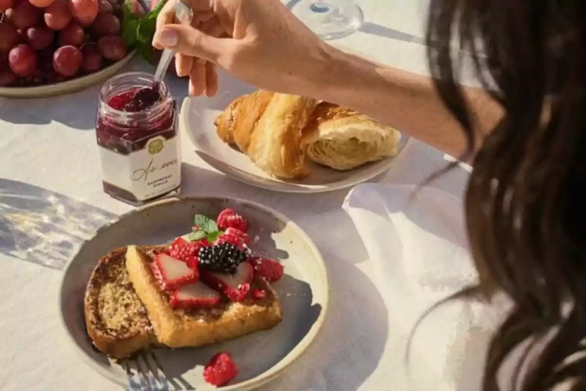 A person spreading jam on toast with fruits and a croissant on the side. A person spreading jam on toast with fruits and a croissant on the side.