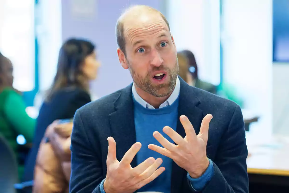 Prince William gestures with his hands while speaking indoors with other people in the background. Prince William gestures with his hands while speaking indoors with other people in the background.