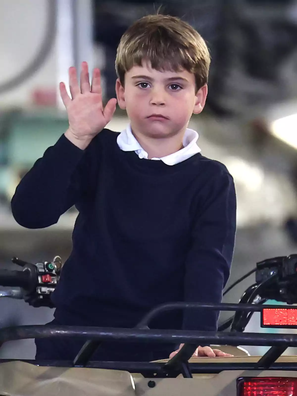 Prince Louis in a dark blue sweater and white jersey raises his hand while standing on a vehicle. Prince Louis in a dark blue sweater and white jersey raises his hand while standing on a vehicle.