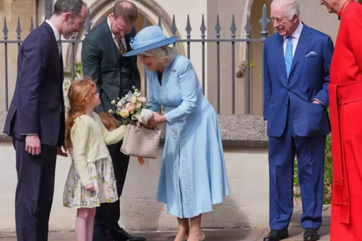 A woman dressed in blue bends down to receive a bouquet of flowers from a little girl while several people watch the scene.