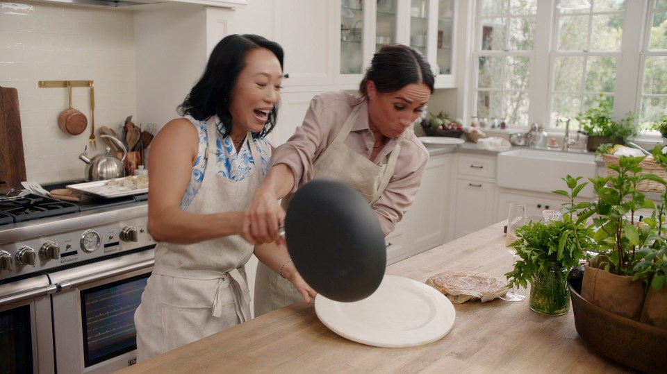 Meghan Markle and Vicky Tsai playfully covering a plate with a pan in a kitchen.