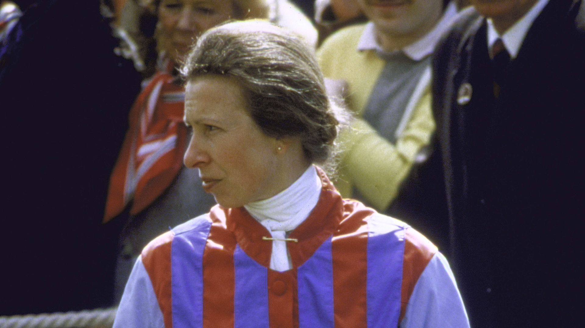 Princess Anne in racing outfit standing amongst others at benefit race in 1985