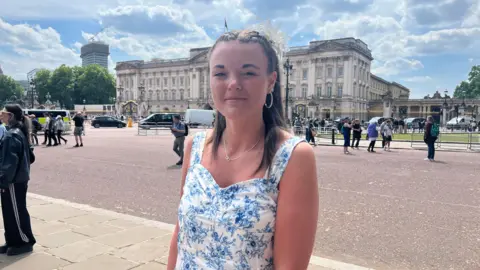 Karl Bird/ BBC A woman with long brown hair wearing a blue and white patterned dress smiling at the camera. She is stood in front of Buckingham Palace. People can be seen gathered or walking in the background. The sky is sunny with white clouds. 