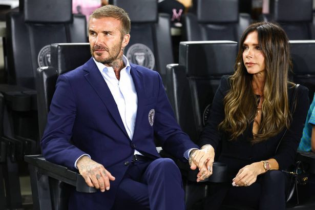 David Beckham and Victoria Beckham, in suits, sat in seats at a football stadium.