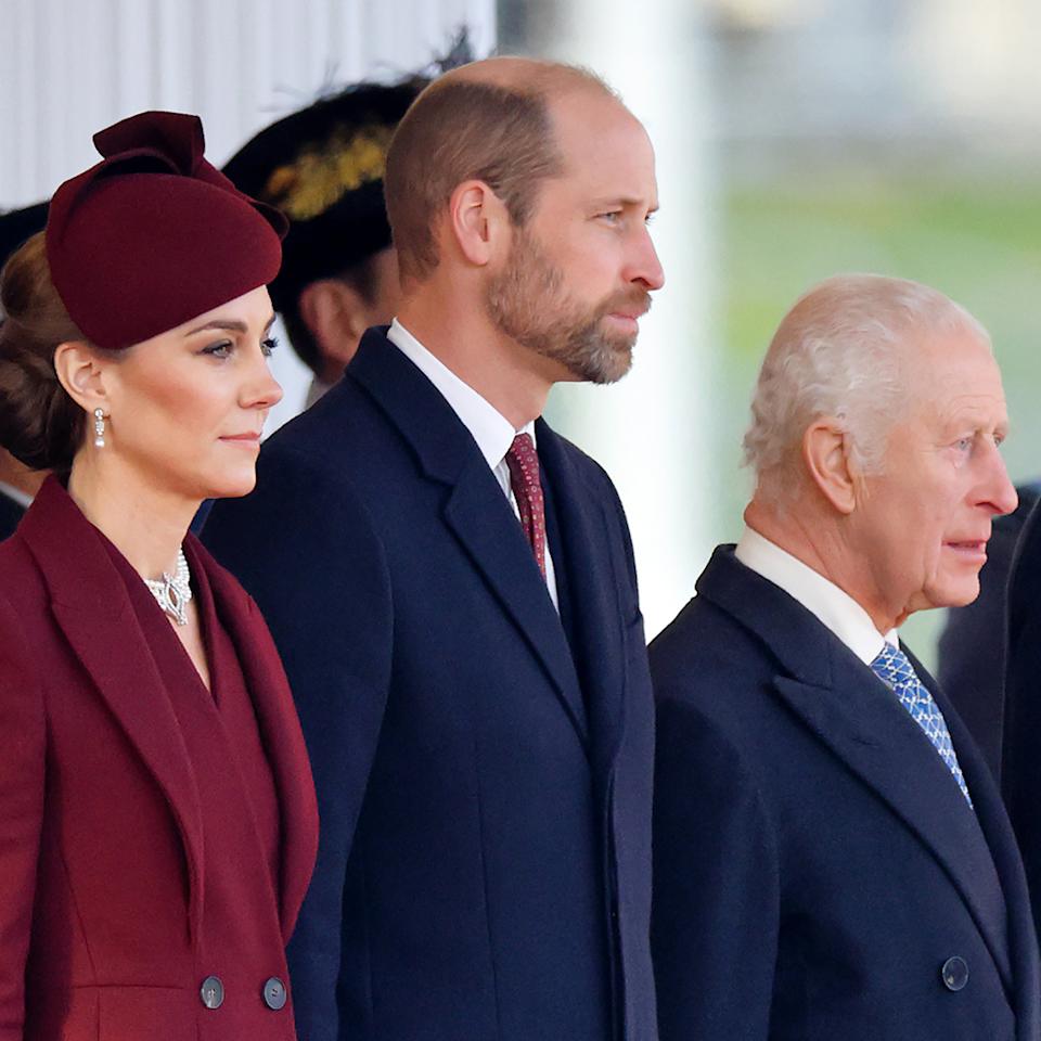 Kate Middleton wears a burgundy coat and matching hat, Prince William wears a navy coat and red tie, and King Charles wears a navy coat with a white shirt and blue tie.