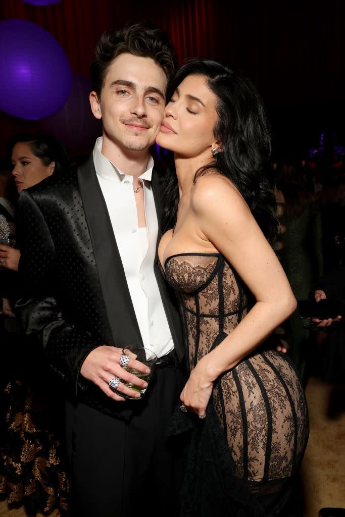 Two individuals at an event, one wearing a black patterned suit, and the other in a strapless lace dress with a corset design