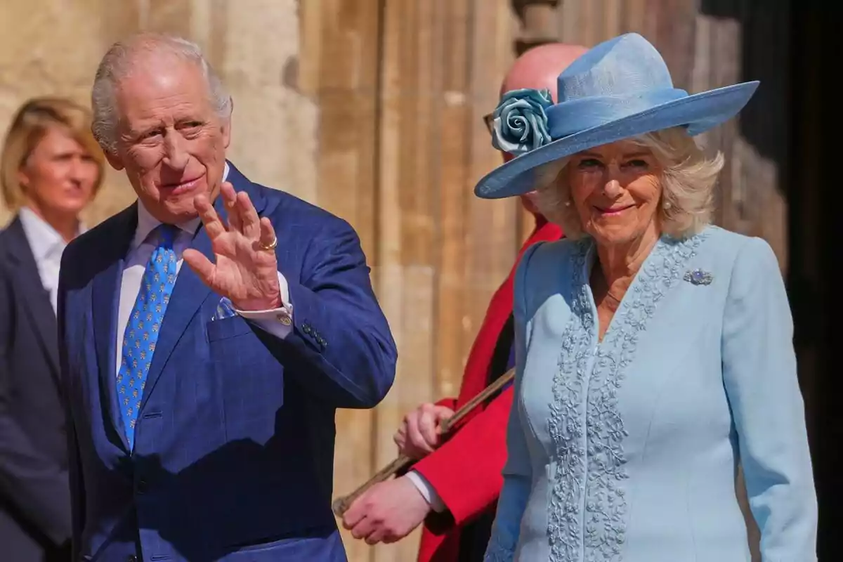 Two elegantly dressed elderly people, one wearing a blue suit with a matching tie, wave as they walk outdoors, the other person is wearing a light blue dress with a hat adorned with a flower.