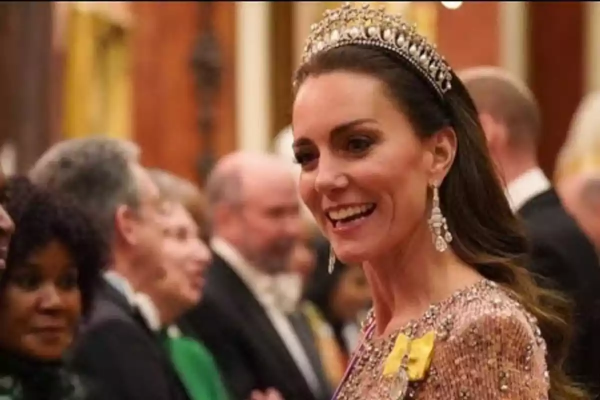 Kate Middleton wearing a tiara and an elegant dress smiles at a formal event surrounded by other people.