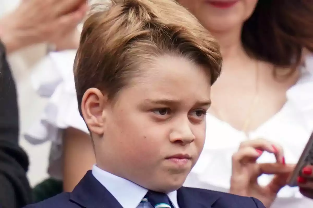 A boy with a serious expression, dressed in a suit and tie, surrounded by people at an event.