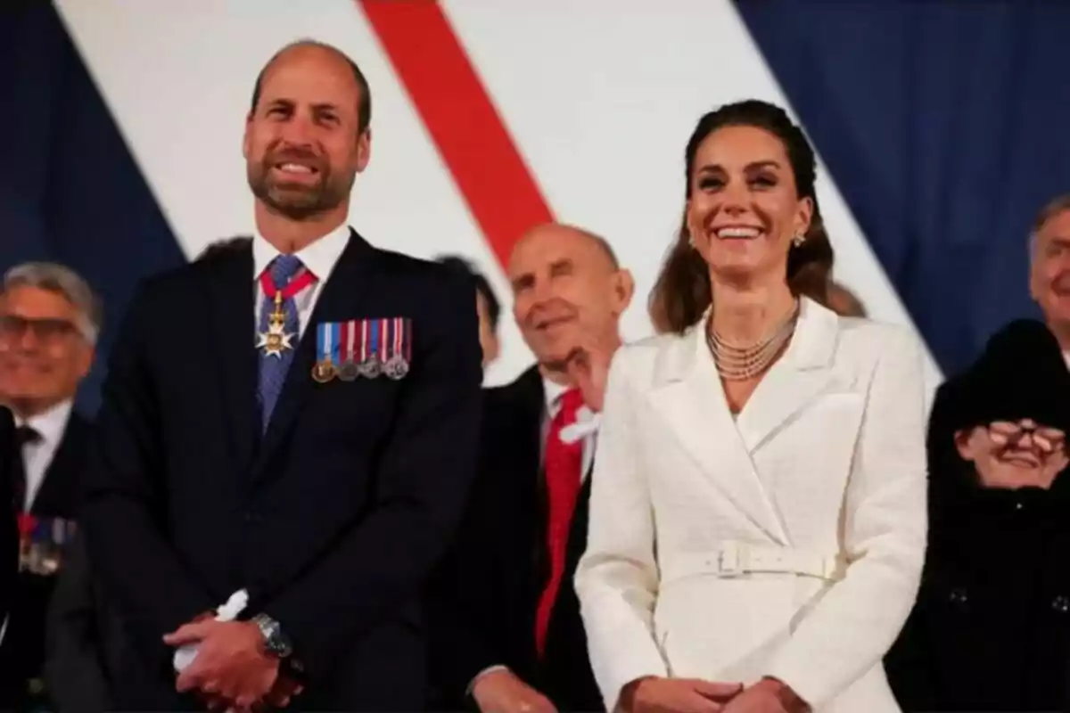 A man and a woman smile while standing at a formal event surrounded by other people.