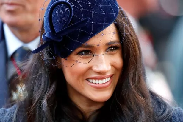Meghan Markle smiling while wearing a navy blue netted fascinator 