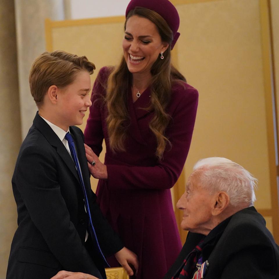 Princess Kate laughing putting her arm on George's shoulder, who is chatting to a WWII veteran.