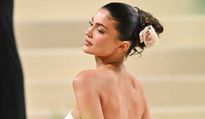 Kylie Jenner on the red carpet wearing an elegant, strapless gown with hair styled up, adorned with a rose accent, looking over her shoulder