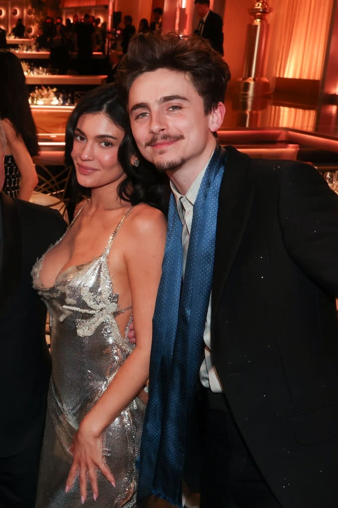 A man and a woman pose together at an event. The woman wears a shimmering gown with embellishments, and the man is in a suit with a scarf