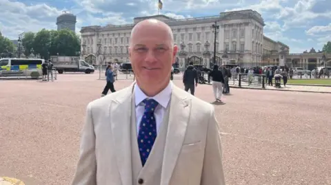 Karl Bird/BBC A man wearing a cream-coloured suit jacket, a cream-coloured waistcoat and a white t-shirt. He is also wearing a blue spotty tie. He is smiling at the camera and is stood in front of Buckingham Palace. People can be seen gathered or walking in the background. The sky is sunny with white clouds. 