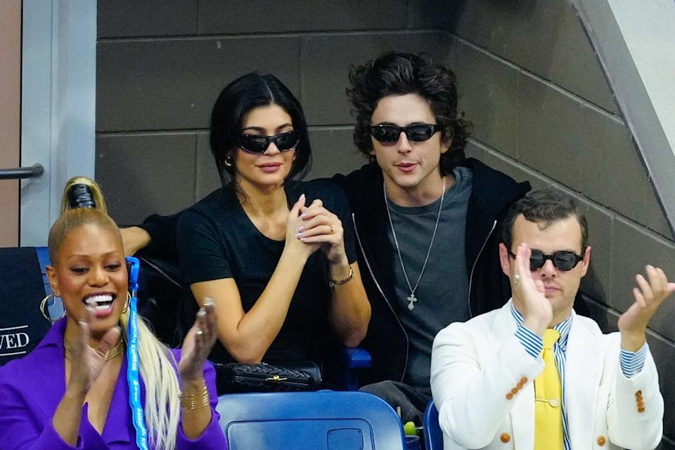 Four people sitting at an event; two in dark chic outfits and sunglasses, another in a purple top with bright earrings, and one in a white jacket clapping