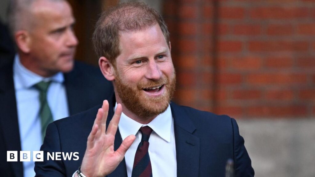 Prince Harry wearing a suit, waving towards a camera.