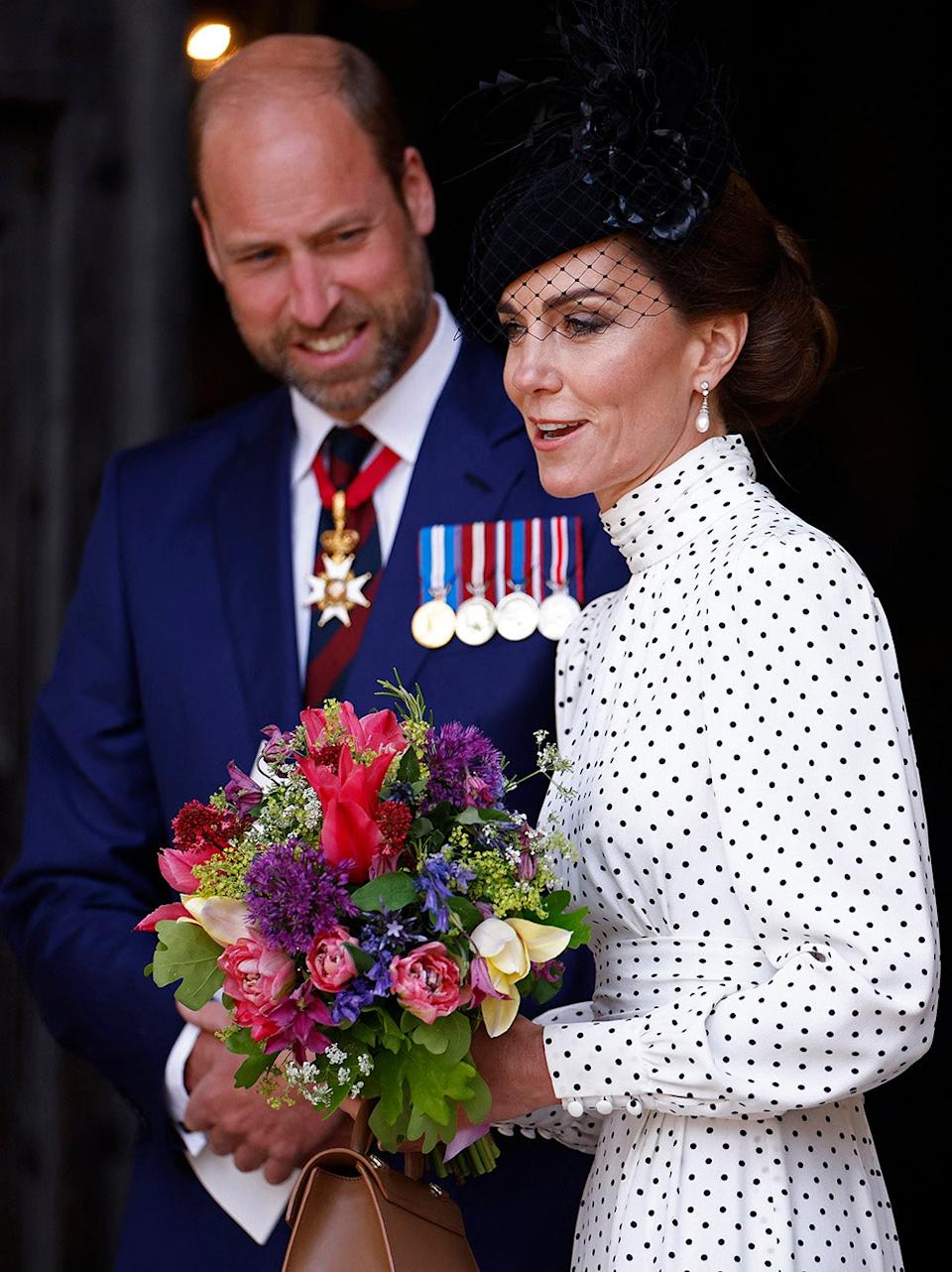 Prince William in a navy suit looking at Kate Middleton holding a bouquet of flowers wearing a polka dot dress and a black fascinator.