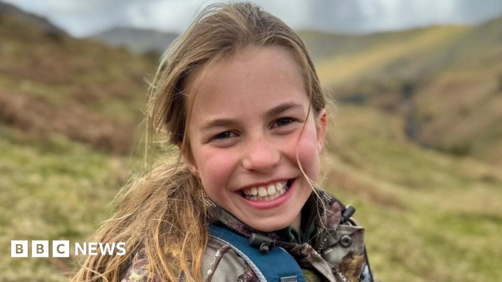 Princess Charlotte smiles to the camera while sitting on a hillside wearing a camoflage jacket and blue backpack