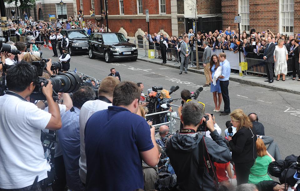 Kate Middleton and Prince William standing in front of a crowd of photographers with baby Prince George