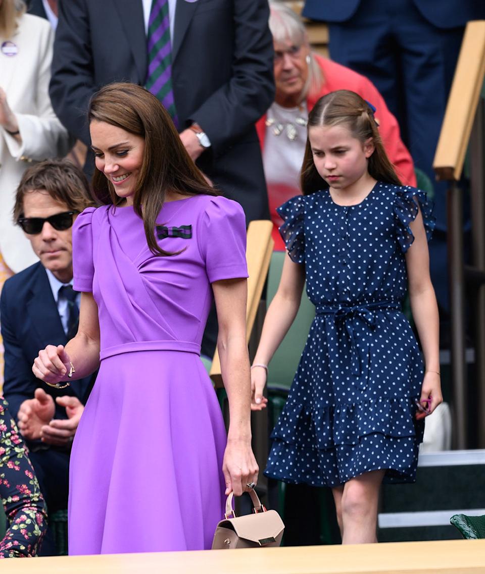 Kate Middleton wearing a bright purple dress walking in front of Princess Charlotte who is wearing a polka dot dress.