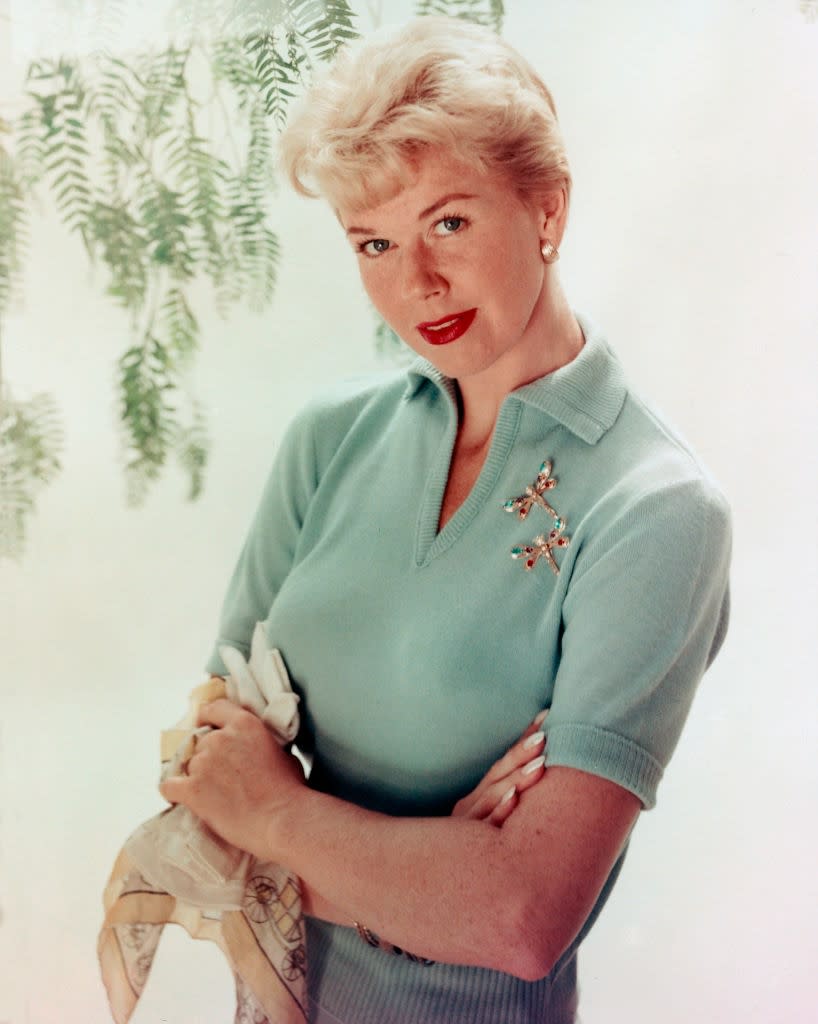 Person in a collared sweater with retro hairstyle and brooch, holding a scarf, poses in front of leafy background