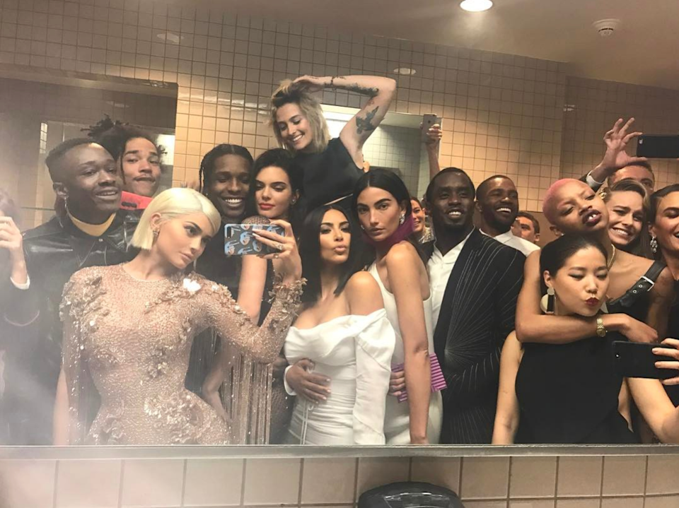 A group of celebrities in stylish outfits take a selfie in a bathroom, with some smiling and others posing
