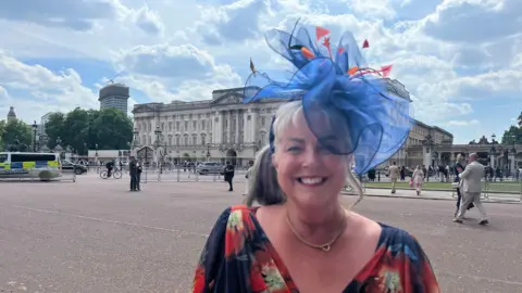 Karl Bird/ BBC A woman wearing a blue red and yellow floral-patterned dress with a blue fascinator on her head. She has blonde hair tied in a ponytail and is also wearing a gold necklace. She is stood in front of Buckingham Palace. People can be seen gathered or walking in the background. The sky is sunny with white clouds. 