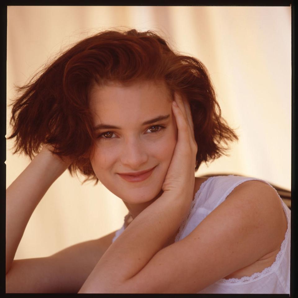 A person with short hair gently resting their head on their hands, smiling at the camera, wearing a lace-trimmed attire