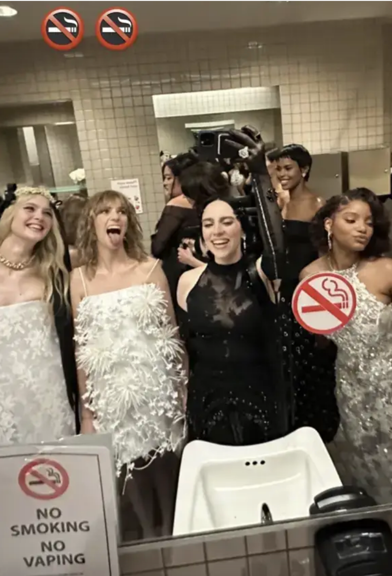 Group of people in a bathroom, wearing elegant dresses, taking a lively photo. Signs indicate no smoking or vaping