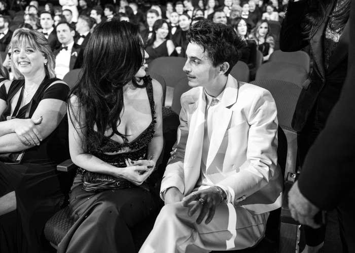 Two people sit at an event, engaging in conversation. One wears a tailored suit and the other a formal dress with a plunging neckline
