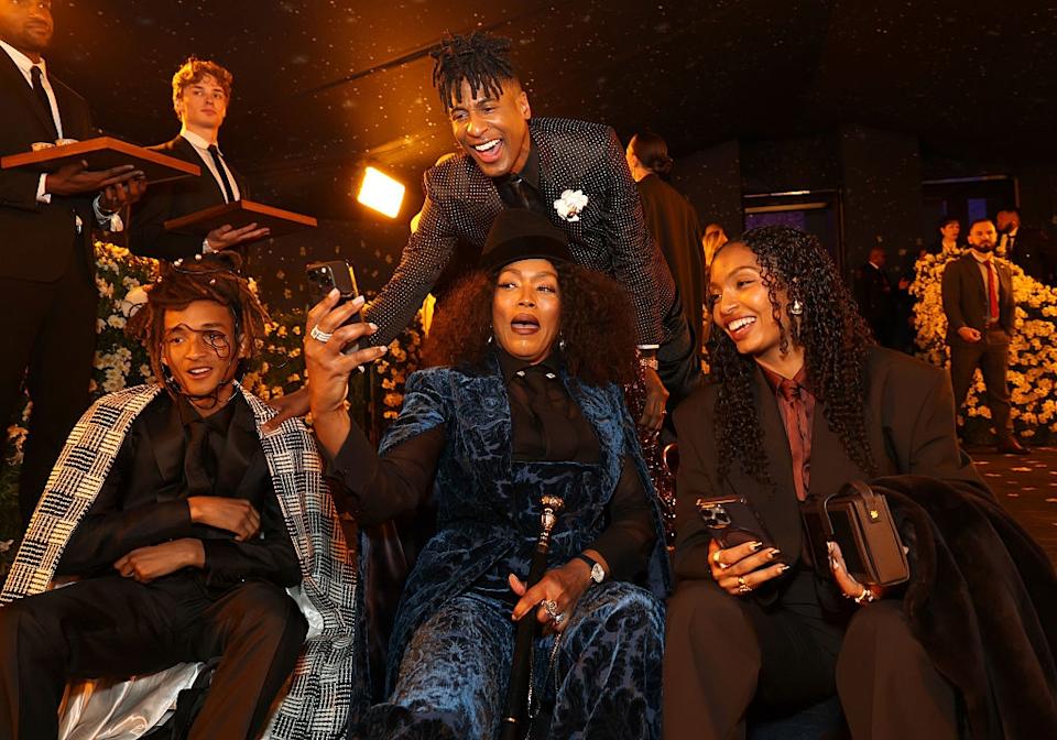 Four people in stylish outfits gather, smiling and taking a group selfie at a glamorous event