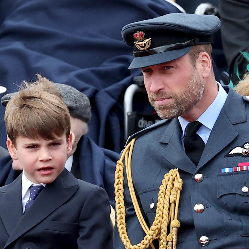  Prince Louis wearing a suit sitting next to Prince William, wearing a military uniform, at a VE Day parade. 