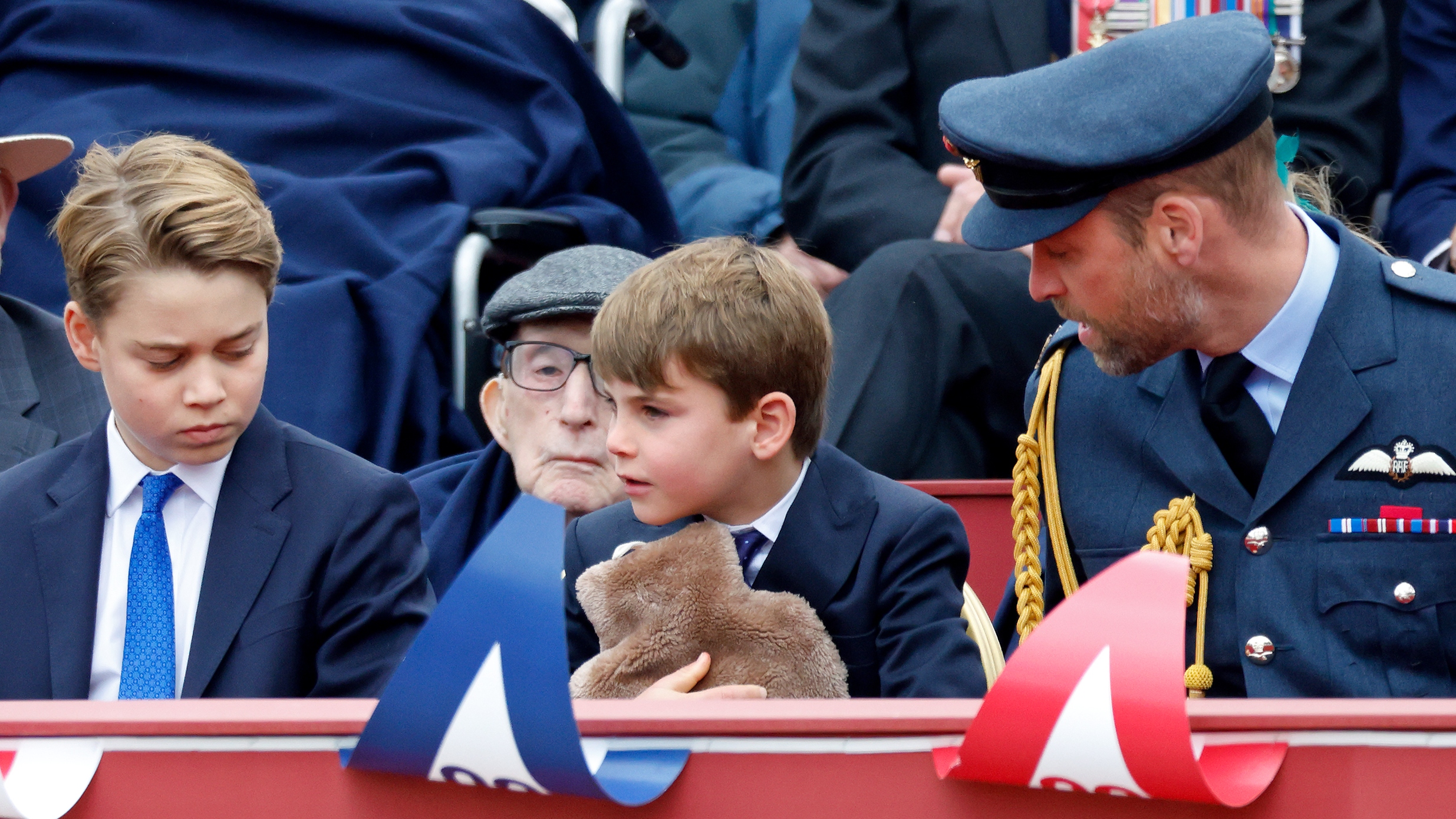 Prince George of Wales and Prince William, Prince of Wales look on as Prince Louis of Wales holds a hot water bottle whilst they attend a military procession
