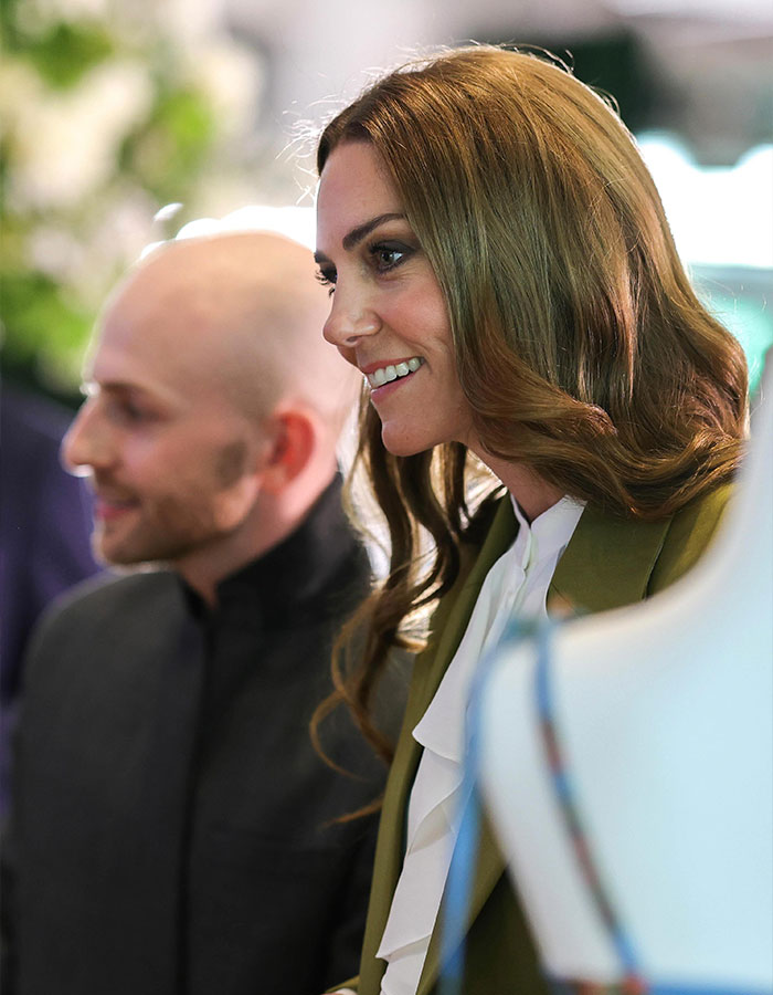Kate Middleton smiling and engaging in conversation with a blurred man in the background at an indoor event. Kate Middleton smiling and engaging in conversation with a blurred man in the background at an indoor event.