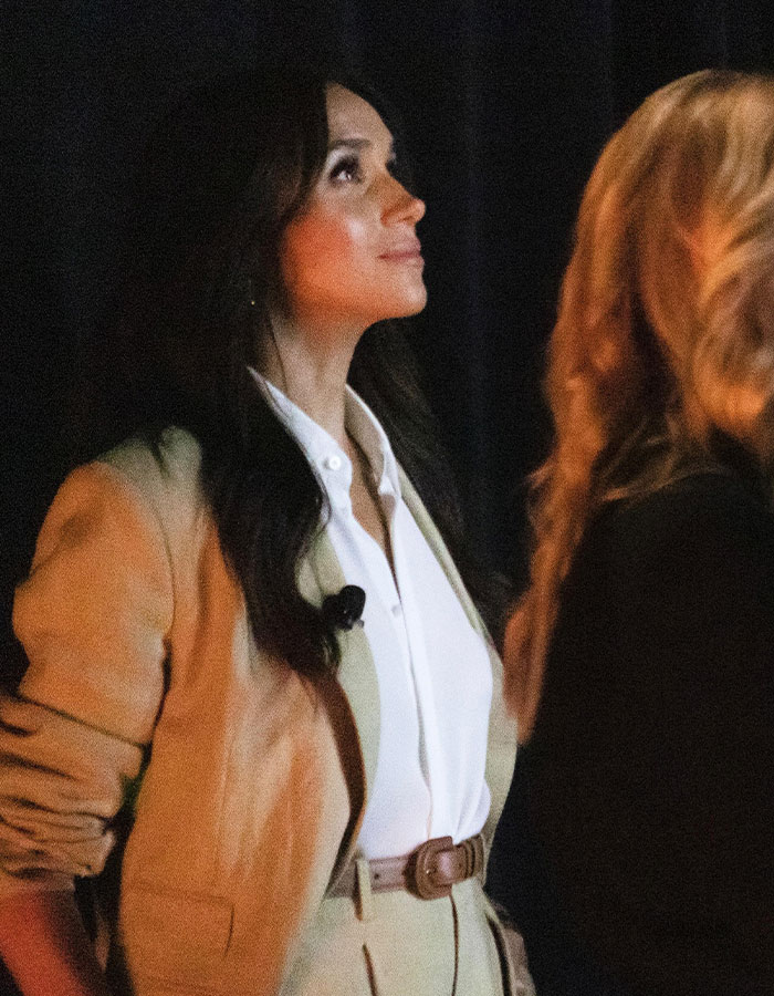 Meghan Markle looking thoughtful wearing a beige suit and white shirt against a dark background during an event. Meghan Markle looking thoughtful wearing a beige suit and white shirt against a dark background during an event.