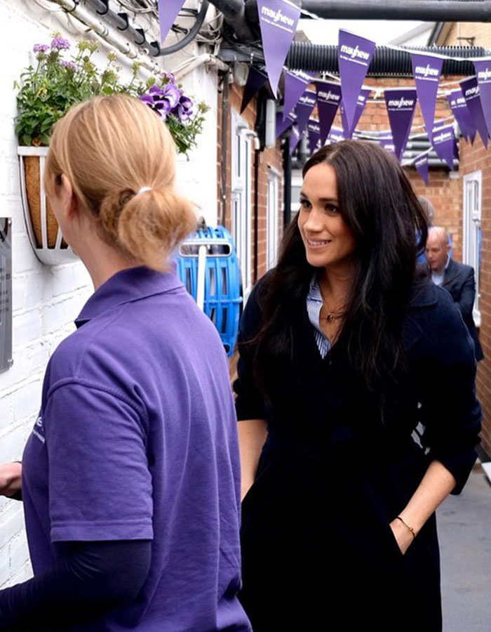 Meghan Markle smiling and talking with a woman in a purple shirt under purple Mayhew banners in a narrow alley. Meghan Markle smiling and talking with a woman in a purple shirt under purple Mayhew banners in a narrow alley.