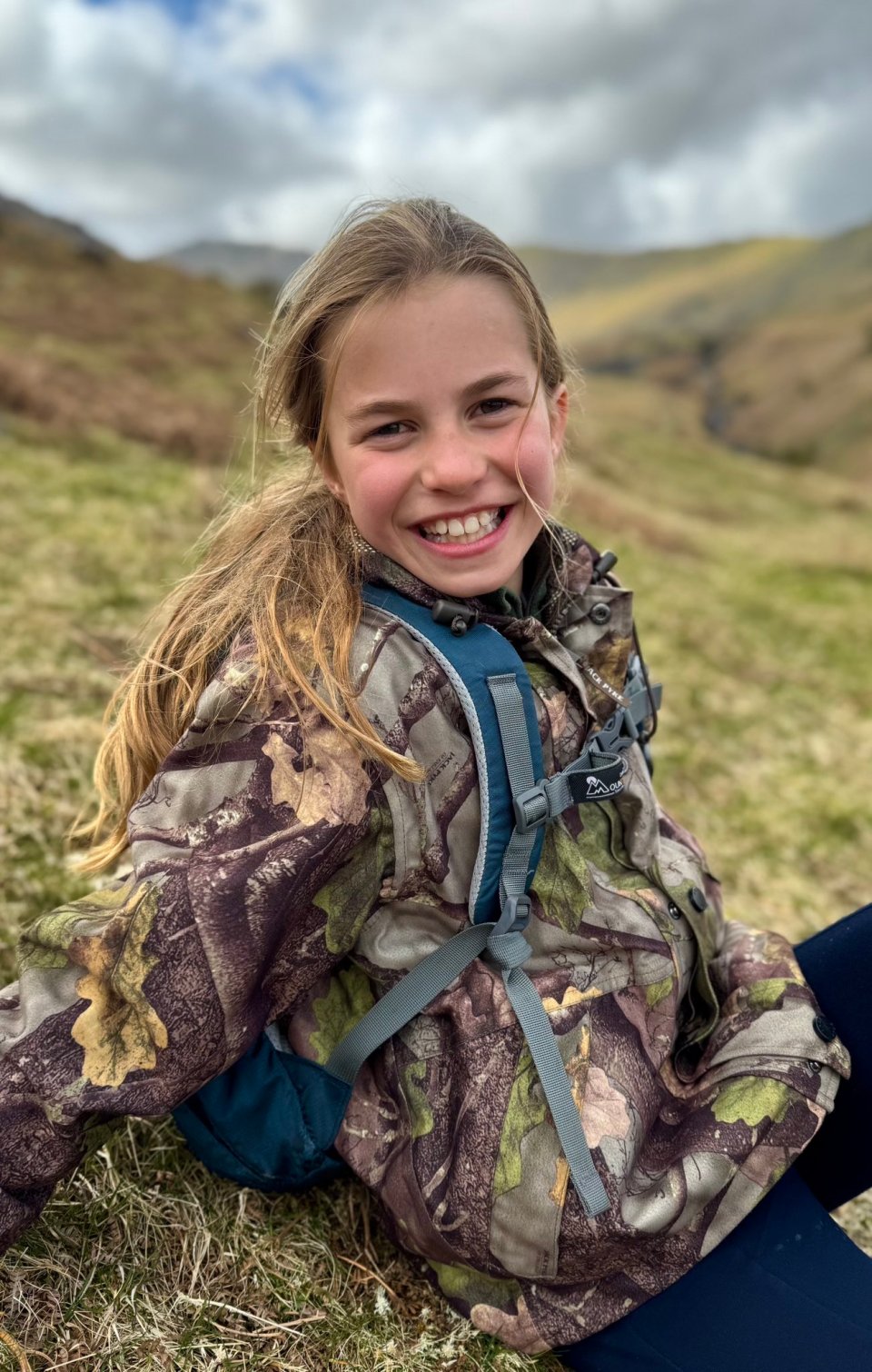 Smiling girl wearing camouflage jacket and backpack outdoors.