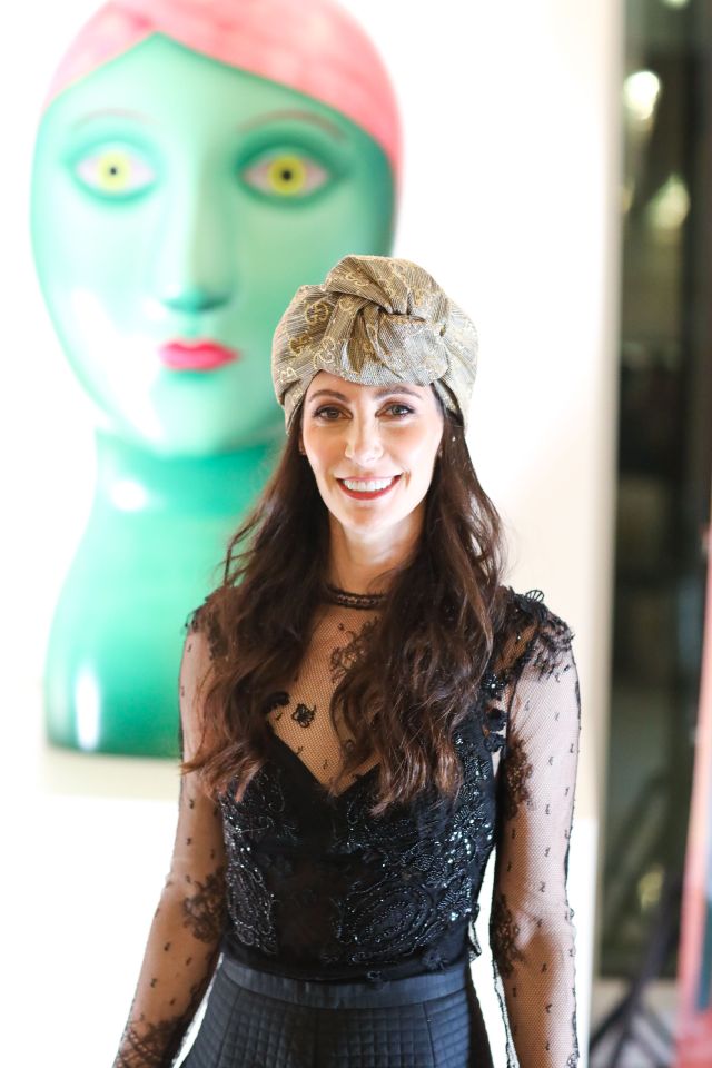 Woman wearing a patterned head wrap and black lace top in front of a large teal sculpture.