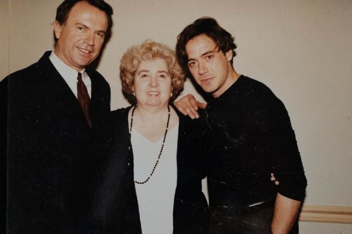 Three people pose together, smiling at the camera. An older woman stands in the middle, flanked by two men—one in a suit and tie, and one in a black shirt with his arm around her shoulder. They are indoors, against a plain wall.