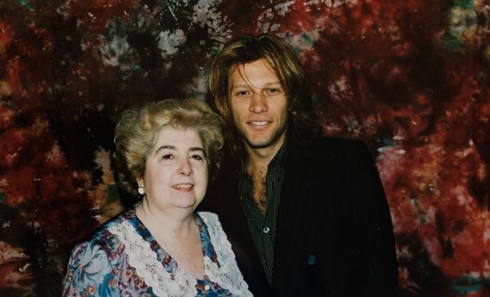 A smiling older woman with curly gray hair and a floral dress stands next to a younger man with long, blondish hair and a dark shirt, both posing in front of a red and black marbled backdrop.