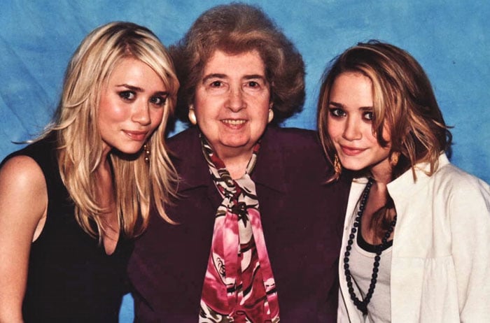 Two young women with blonde hair stand on either side of an older woman with gray hair. All three are smiling in front of a blue backdrop. The older woman wears a patterned scarf and suit jacket.
