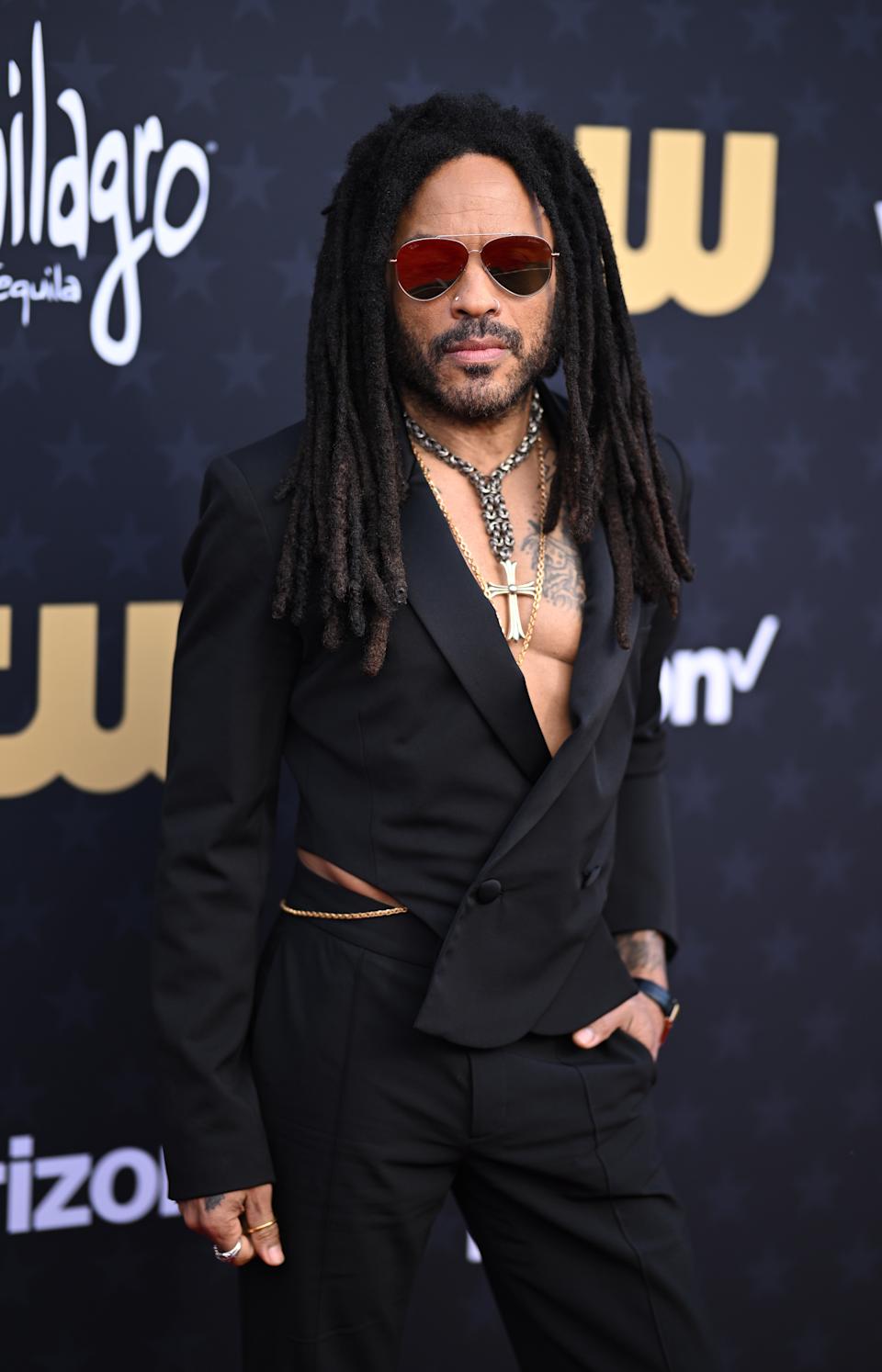 Lenny Kravitz wearing a stylish, unbuttoned black suit and sunglasses on a red carpet