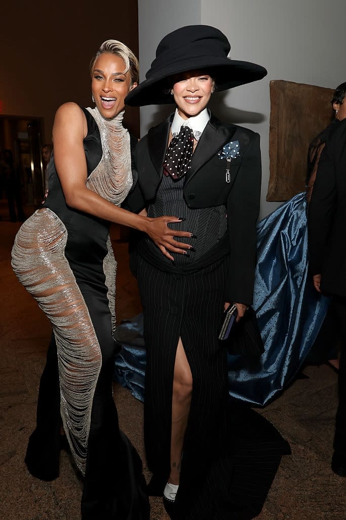 Two individuals pose stylishly; one in a sheer-patterned dress, the other in a tailored suit with a large hat, exuding elegance and confidence