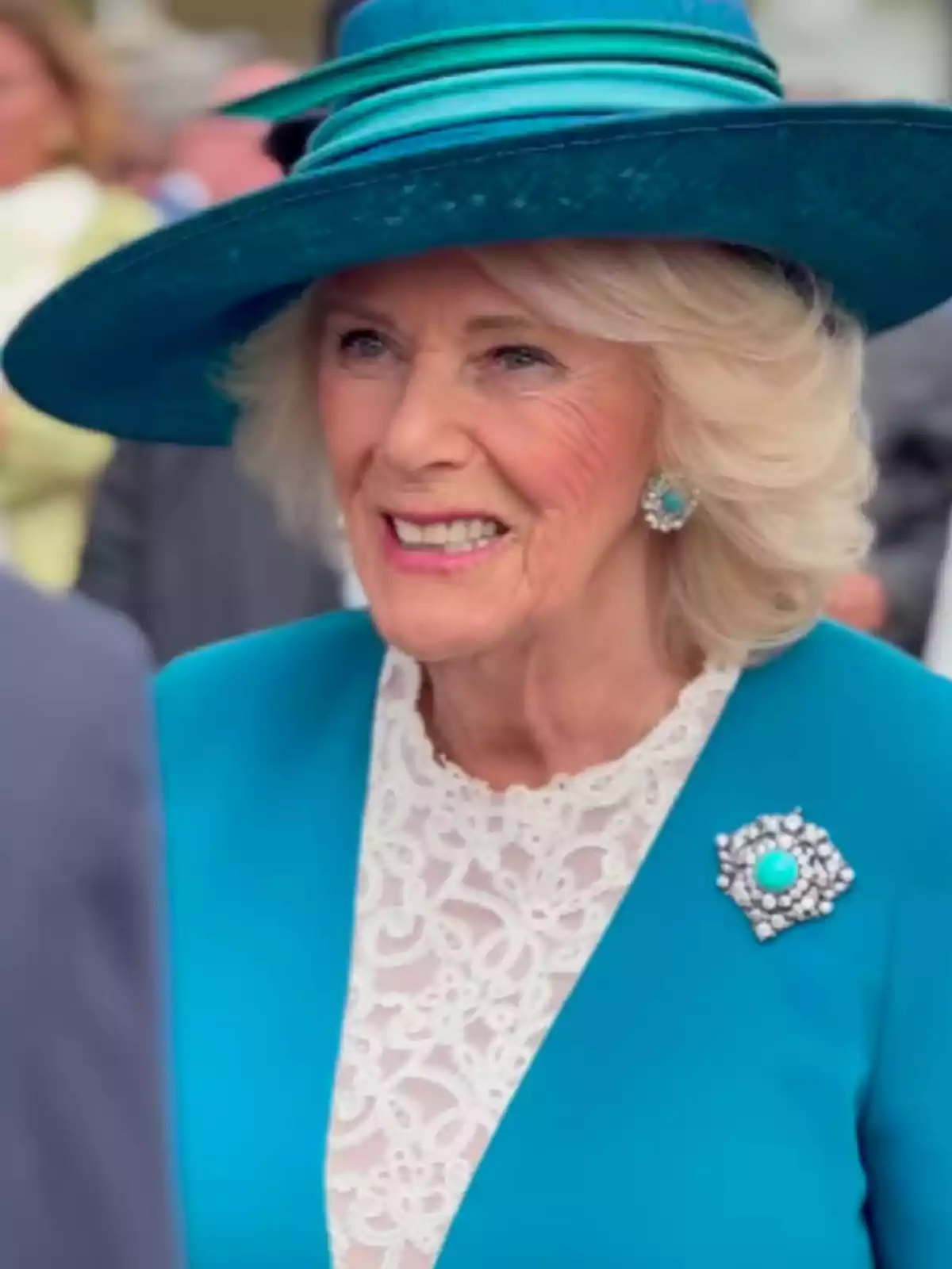 A smiling woman with a blue hat and a matching brooch in her elegant outfit.