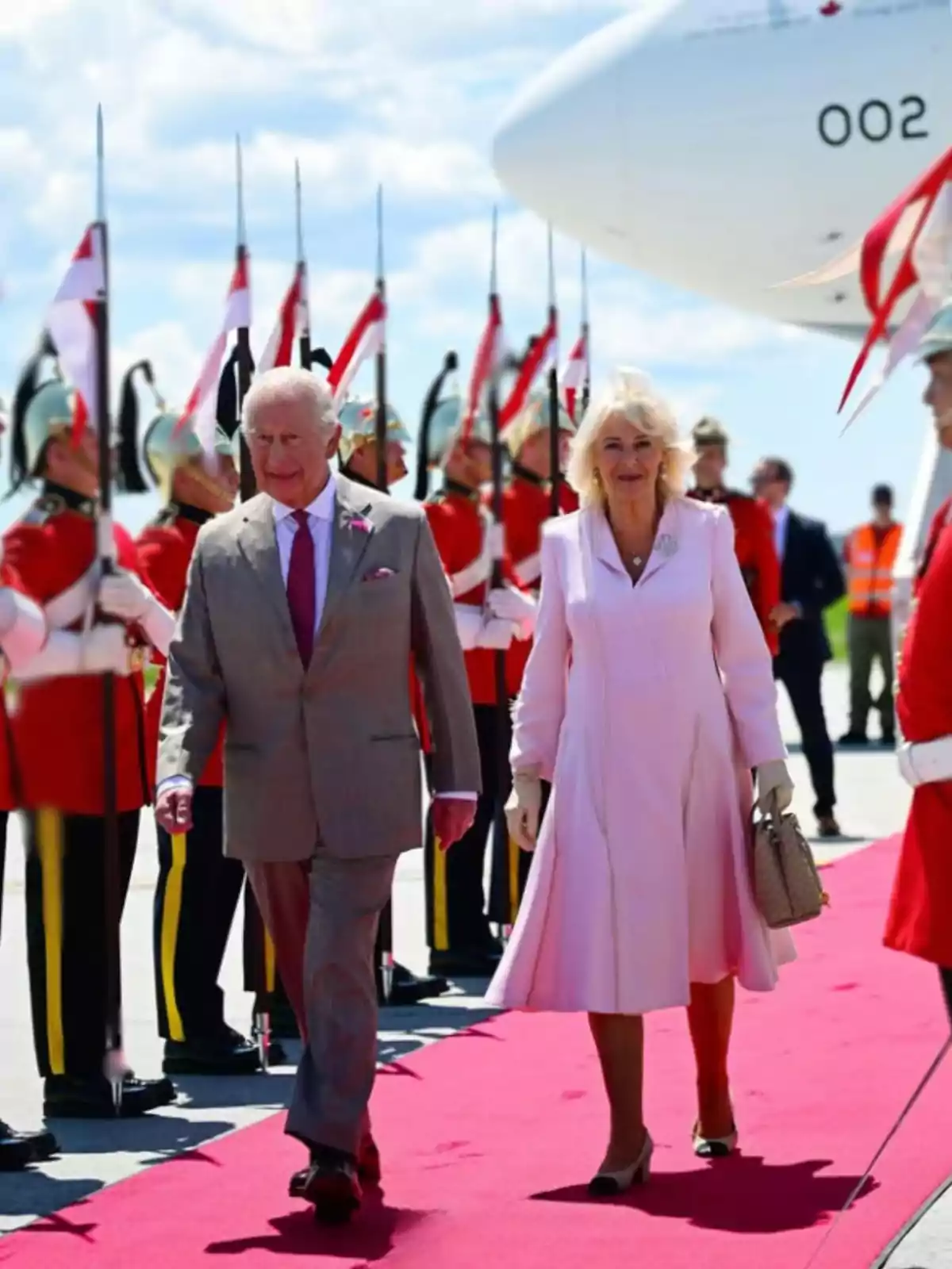 An elegantly dressed couple walks down a red carpet while soldiers in dress uniforms and carrying spears form a guard of honor next to a white airplane.
